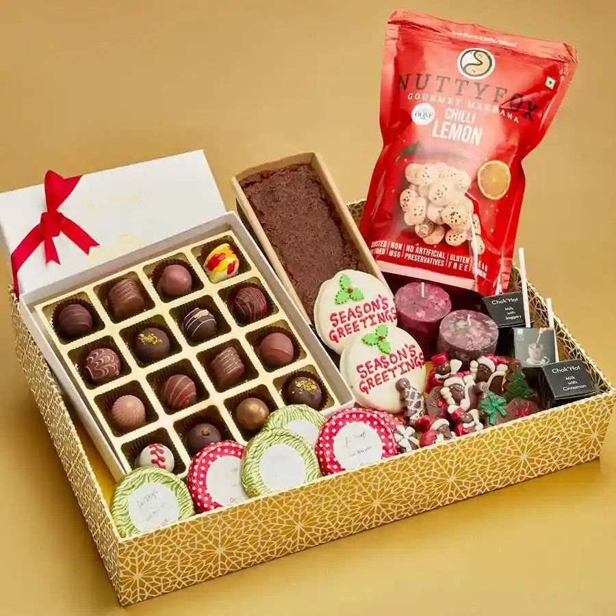 Holiday Indulgence Chocolate N Cake Box featuring a Christmas plum cake, Belgian pralines, chocolate-coated biscuits, chocolate-dipped cookies, Chok Hot sticks, assorted Christmas-shaped chocolates, flavorful makhana, and Christmas candles.