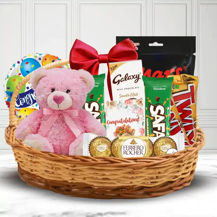 Exquisite gift basket featuring chocolates, a teddy bear, a congratulations card, and a vibrant balloon, all beautifully arranged in a wicker basket with a handle and bow.