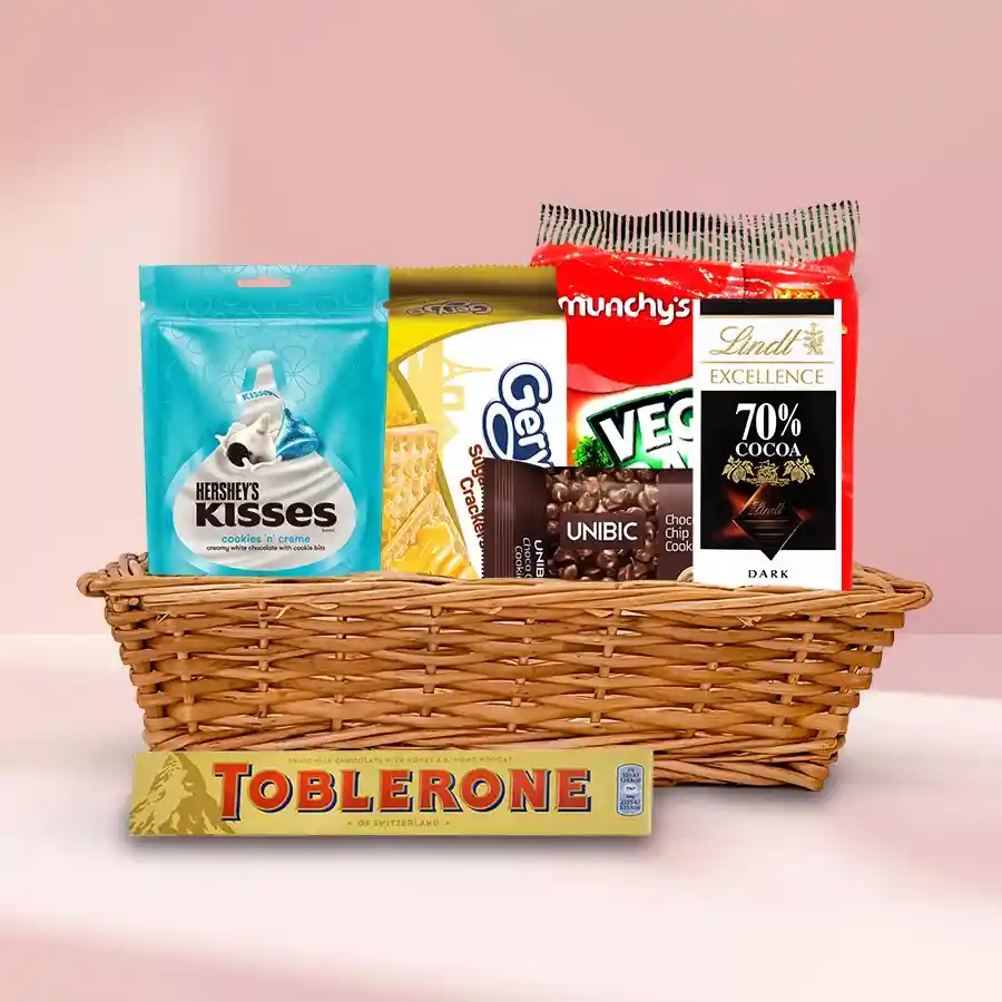 A beautifully arranged small basket filled with various chocolate treats including Lindt and Toblerone bars, Hershey's Kisses, Gery and Munchys crackers, and Unibic Choco Chip Cookies.