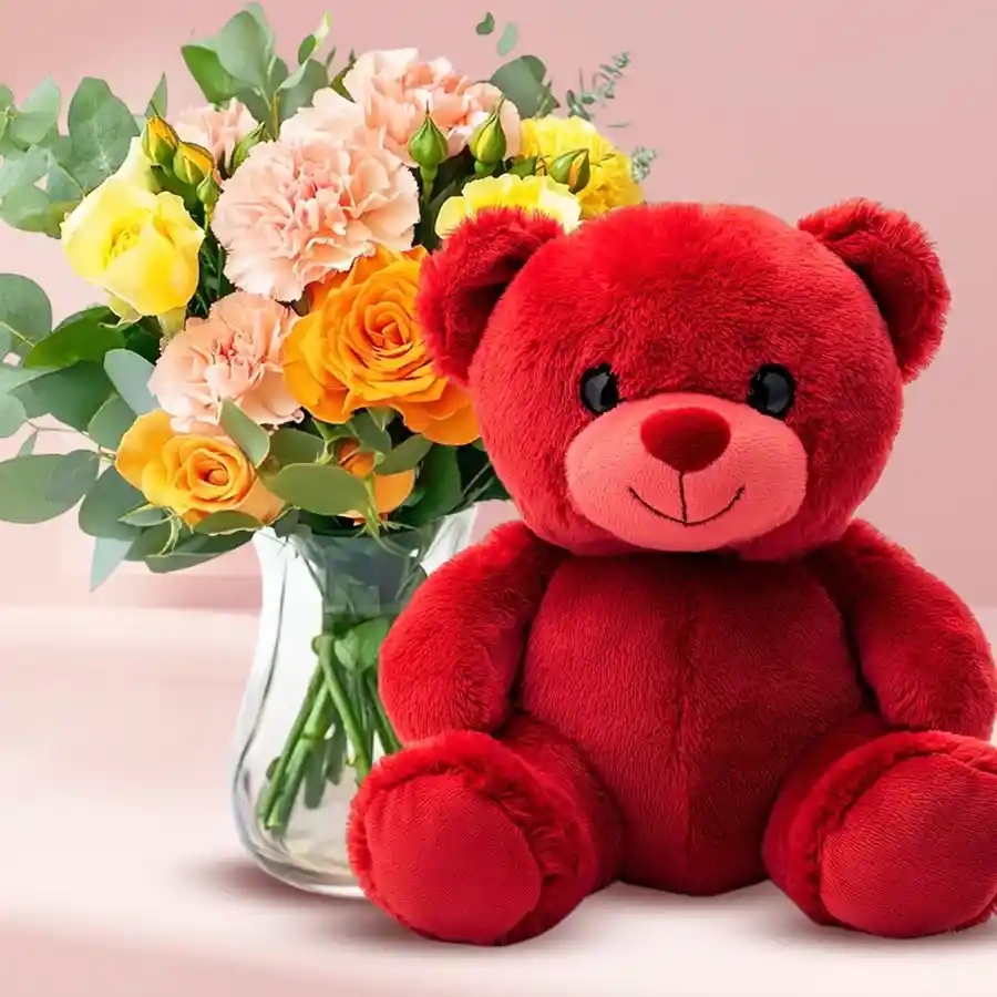 A charming vase with orange roses, pink carnations, yellow roses, and a soft red teddy bear.