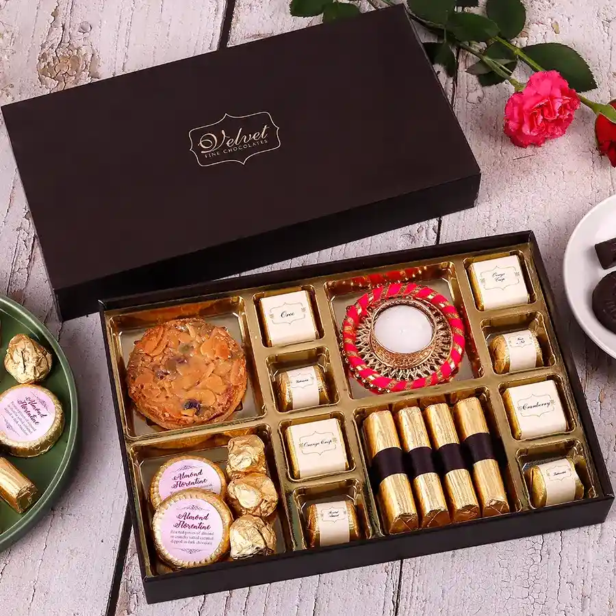 A beautifully arranged box of festive sweets including Florentine cookies, almond chocolate florentines, assorted gourmet chocolates, chocochip cookies, granola logs, and a decorative diya on top.