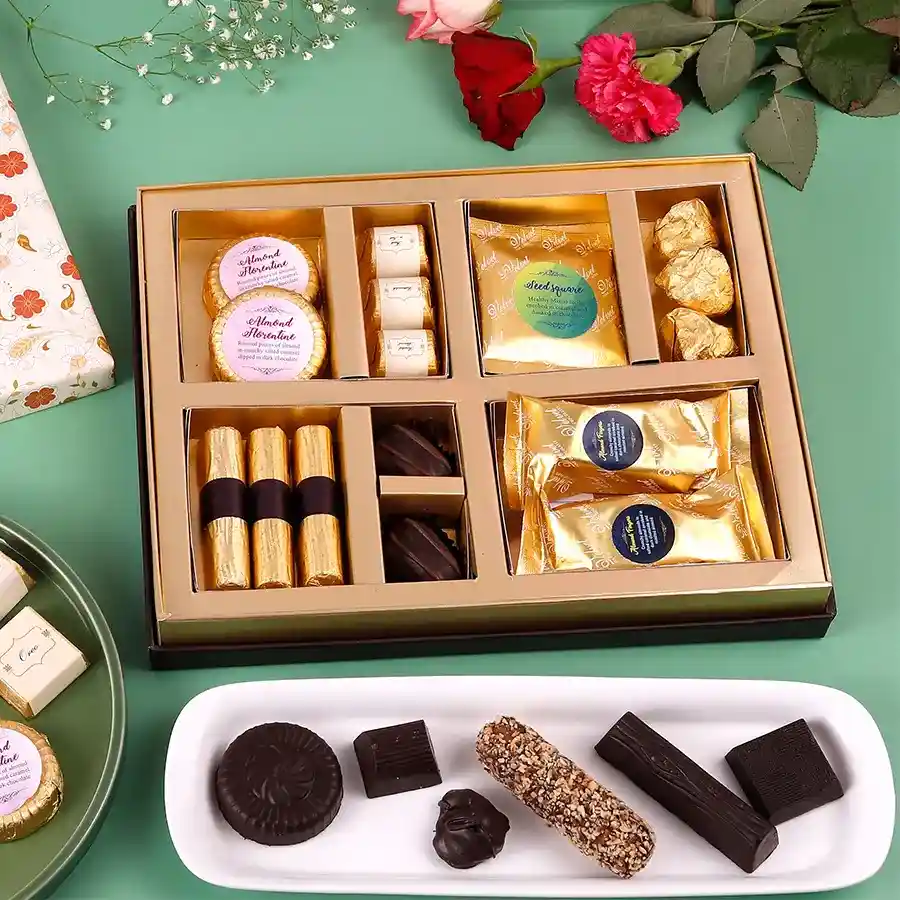 A vibrant box filled with assorted festive treats including almond fingers, butterscotch chocolates, granola logs, almond chocolate rocks, seeds square, assorted chocolates, and almond florentines, set against a decorative background for Diwali celebrations.