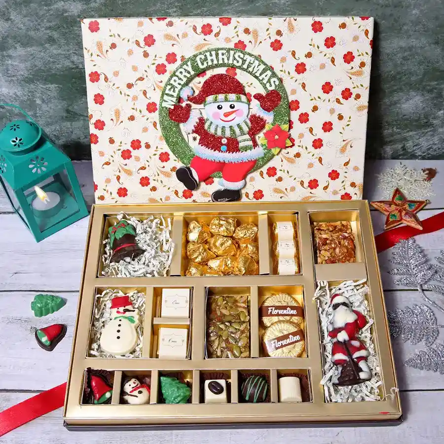 A beautifully decorated floral box filled with assorted chocolates, including a chocolate snowman, a festive bell, almond rocks, almond squares, and a chocolate Santa, perfect for Christmas.
