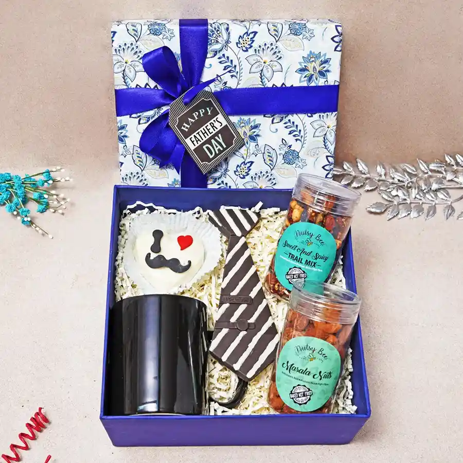 A gift box containing sweet and spicy trail mix, crunchy masala nuts, a chocolate tie, a hot chocolate pinata, and a stylish mug, ideal for Father's Day celebration.