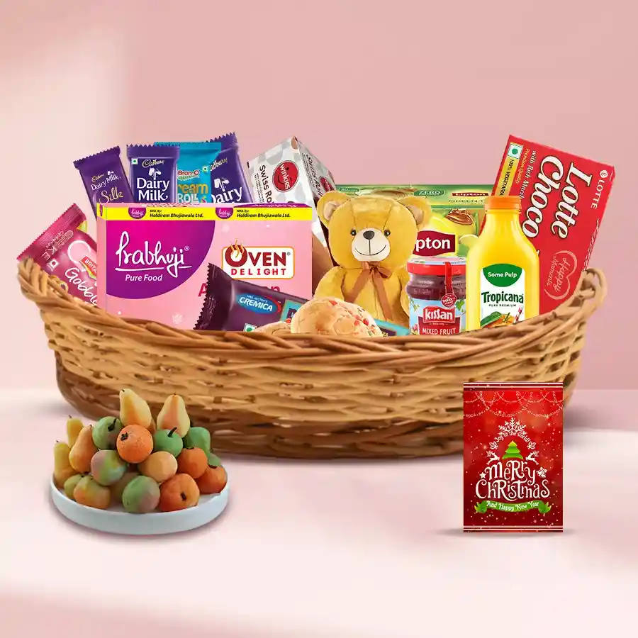 A festive cane basket filled with a variety of holiday treats including fruit cake, cookies, jams, chocolates, and a teddy bear, accompanied by a Christmas card.