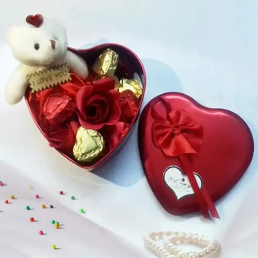 Heart-shaped box filled with handmade chocolates, a small teddy bear, and three fresh roses arranged together for a romantic gift.