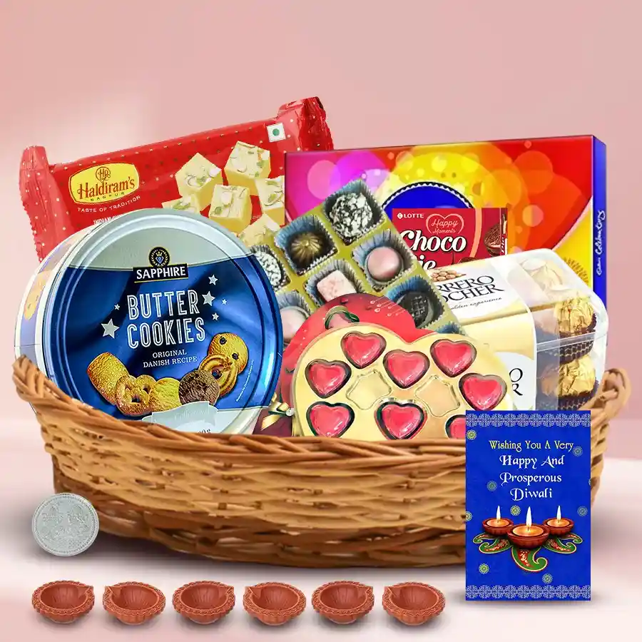 Gift basket filled with assorted chocolates, cookies, Soan Papdi, decorative diyas, an antique silver coin, and a greetings card, arranged in a cane basket for Diwali celebrations.