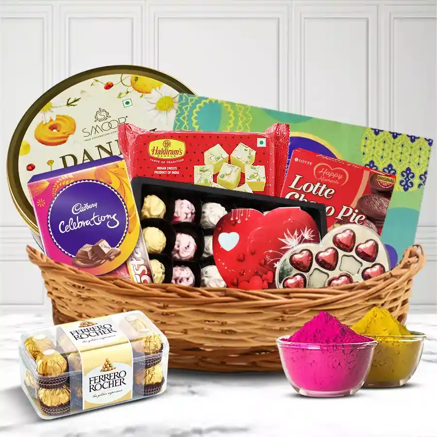 A beautifully arranged cane basket filled with red heart chocolates, assorted homemade treats, Ferrero Rocher, Cadbury Celebration packs, Danish butter cookies, Soan Papdi, Chocopie, and vibrant herbal gulal for Holi celebrations.