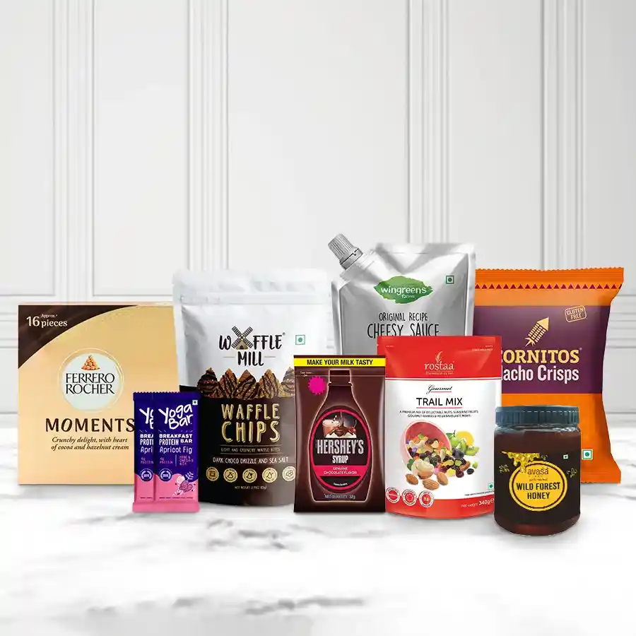 Gift hamper filled with sweet and savory treats for Christmas, including chocolate chips, nachos, trail mix, protein bars, honey, cheese sauce, and chocolate syrup.