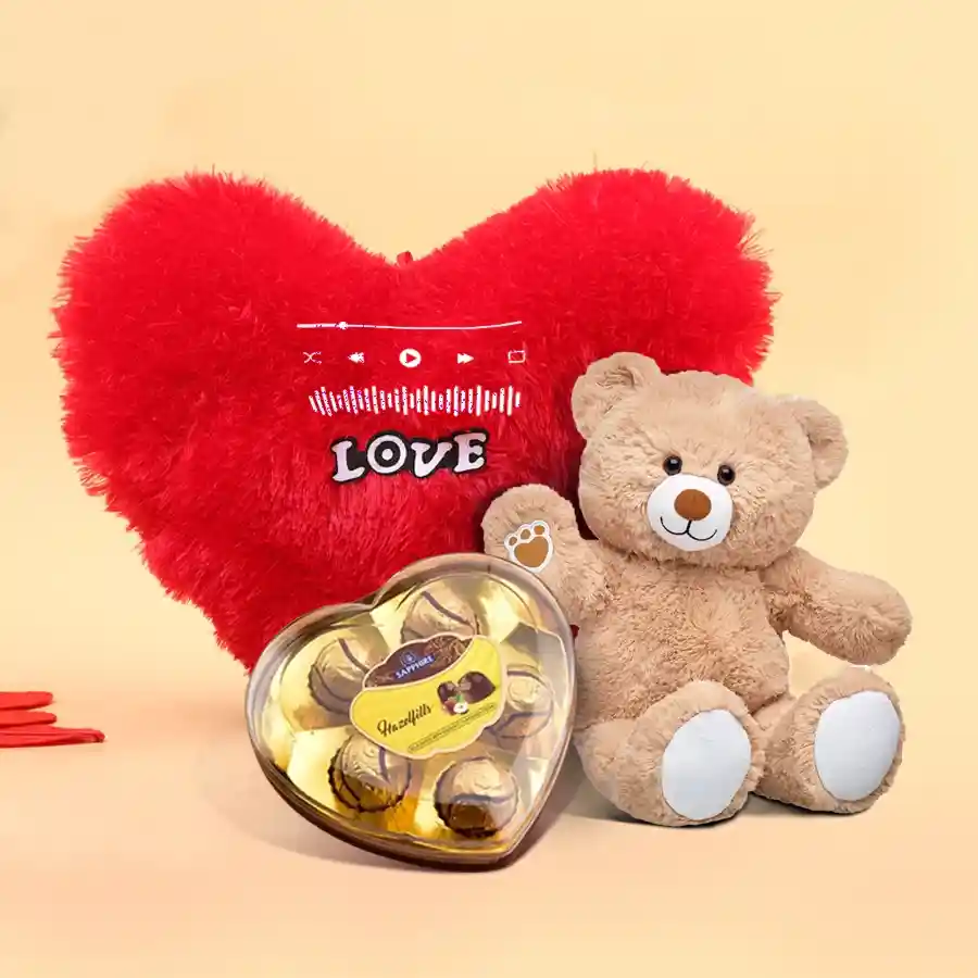 Heart-shaped cushion with a teddy bear and a box of heart-shaped chocolates arranged for a Valentine's Day gift.