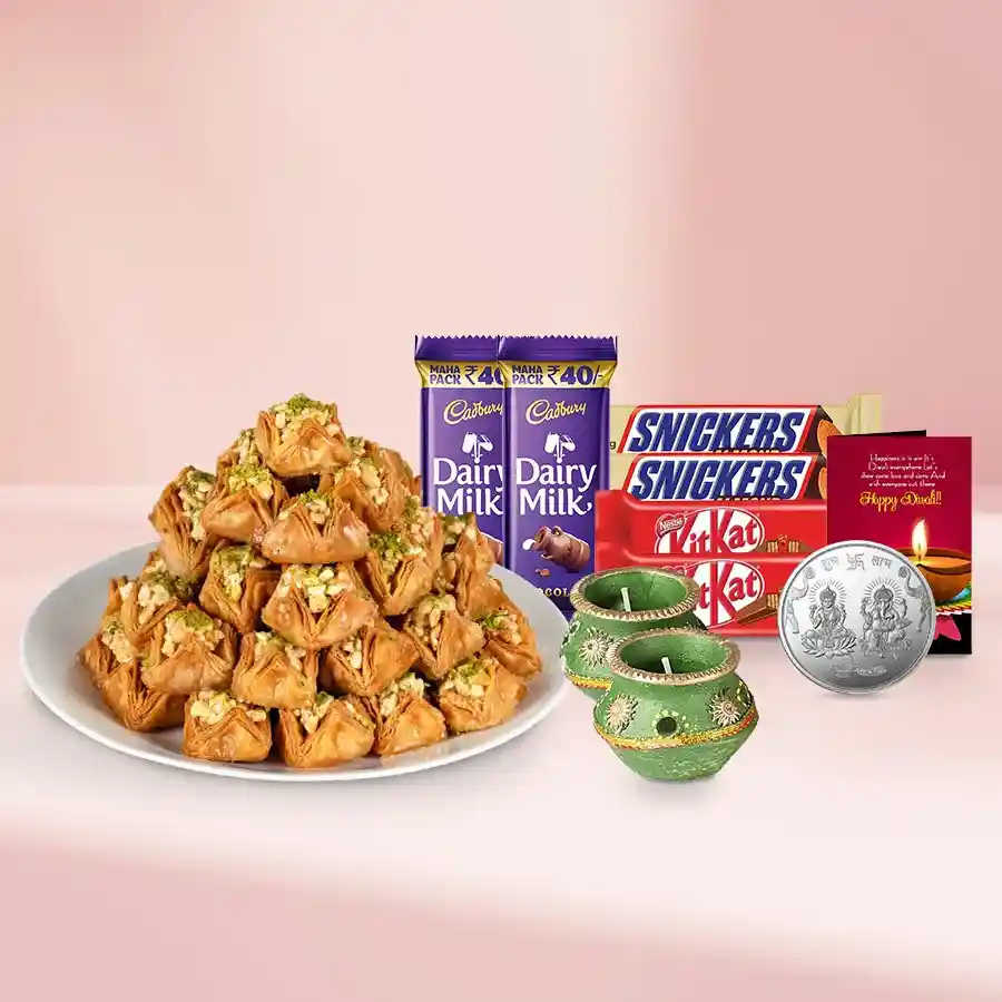 A decorative basket filled with Pyramid Baklava, Snickers, Cadbury Dairy Milk, KitKat bars, festive wax diyas, and a silver Ganesh Laxmi coin, topped with a Diwali card.