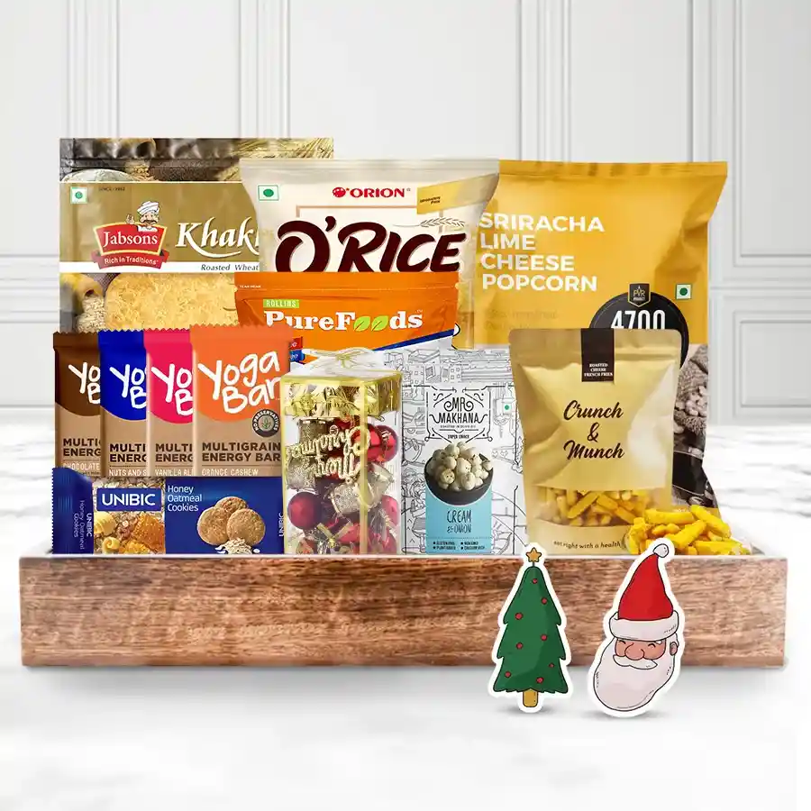 A festive food tray filled with various snacks including Yoga Bars, oatmeal cookies, lime cheese popcorn, fox nuts, banana chips, jeera khakhra, French fries, and rice crackers, adorned with Christmas decorations and wrapped in a reusable gift basket.