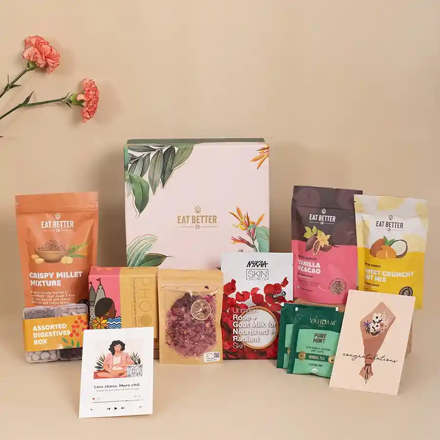 Nature printed gift box filled with indulgent treats including vanilla and cacao laddoos, nut mix, herbal teas, foot salts, face mask, incense cones, millet mix, and digestives.