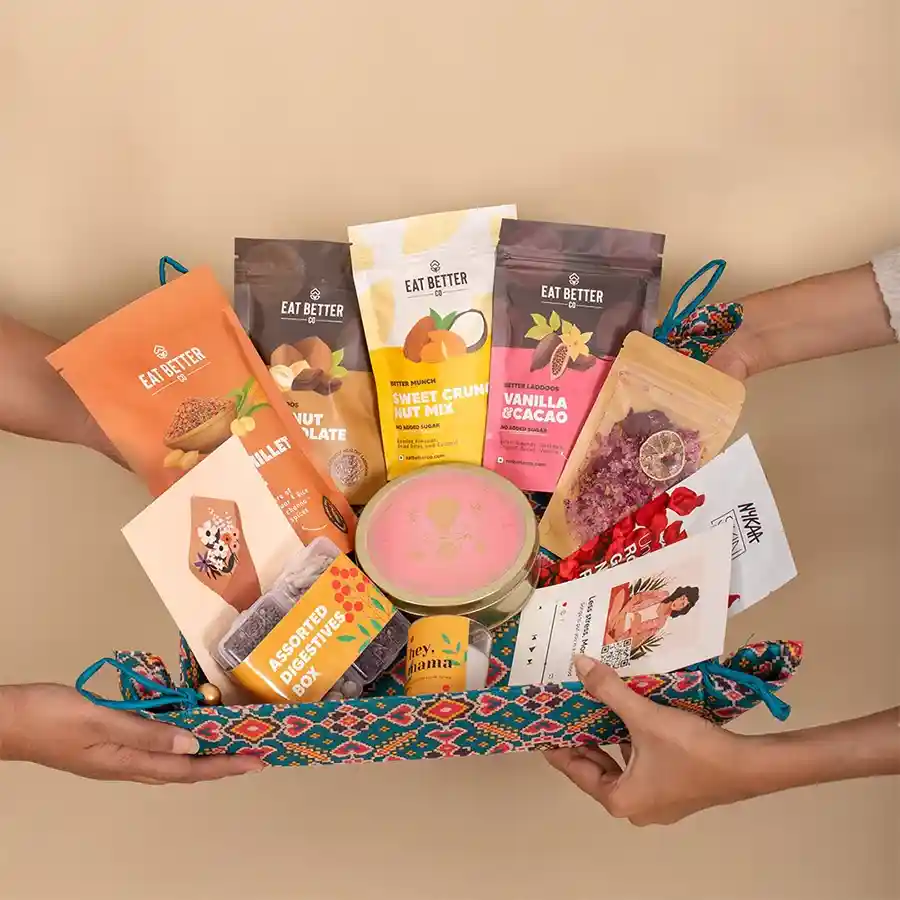 Delectable goodies arranged in a colorful Patola art tray, including sweet treats, nut mix, tea, a candle, foot salts, face masks, digestives, and a curated playlist.
