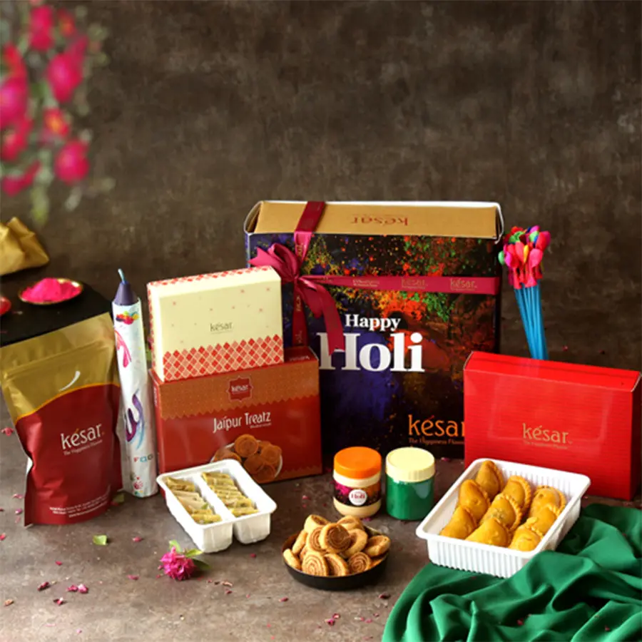 Holi treat hamper featuring Kesar Gujiya, Bhakarwadi, Thandai Katli, crunchy peanuts, gulal, chandan, water balloons, and a color bomb, arranged in a colorful box with a Holi greeting card.