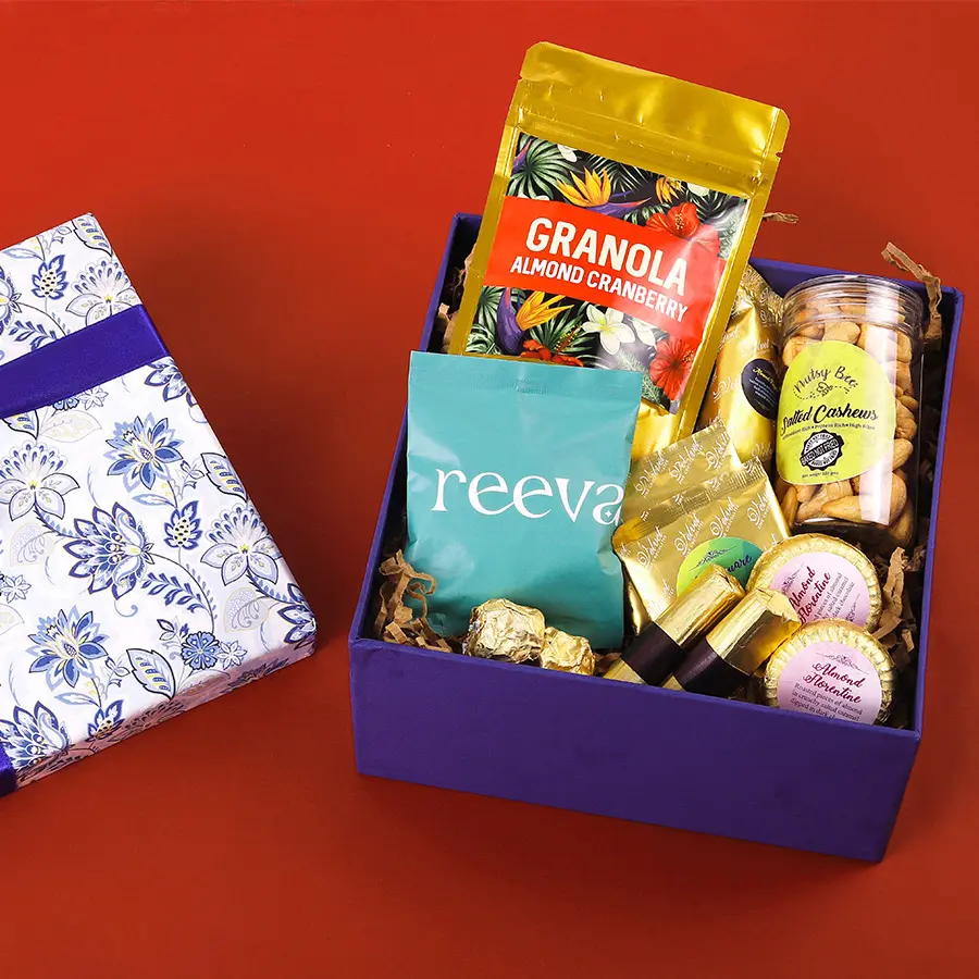 An assortment of gourmet snacks including Almond Cranberry Granola, Almond Chocolate Fingers, Salted Cashews, Chocochip Cookies, Seeds Squares, Almond Chocolate Florentines, Rocks, and Chocolate Logs presented in an elegant package.