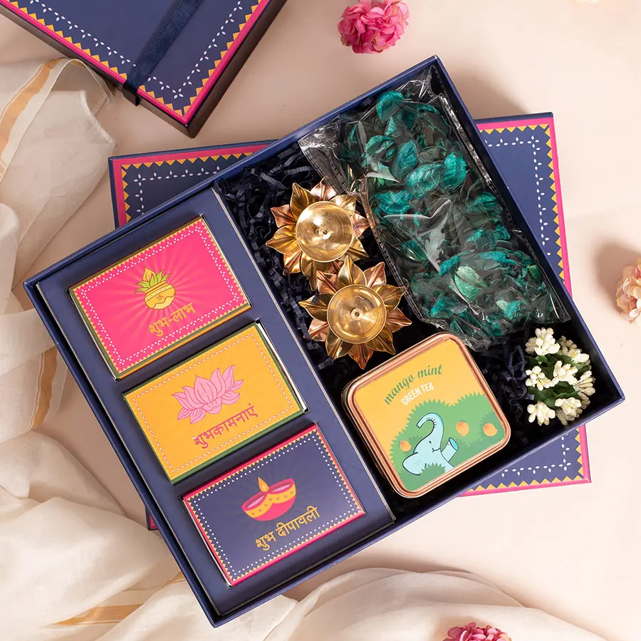 Gleaming Diwali Gift Selection featuring a reusable box filled with premium almonds, cashews, pistachios, two brass diyas, Mango Mint Tea, and green potpourri