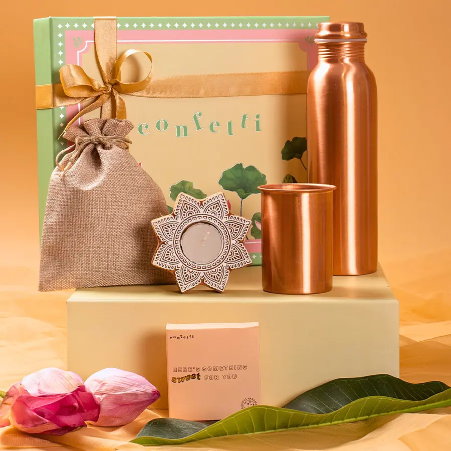 Handcrafted Delights Diwali Treats Box featuring premium dry fruits in a jute potli, an elegant copper glass and bottle, a wooden candle, handmade chocochip cookies, and a green lotus box.