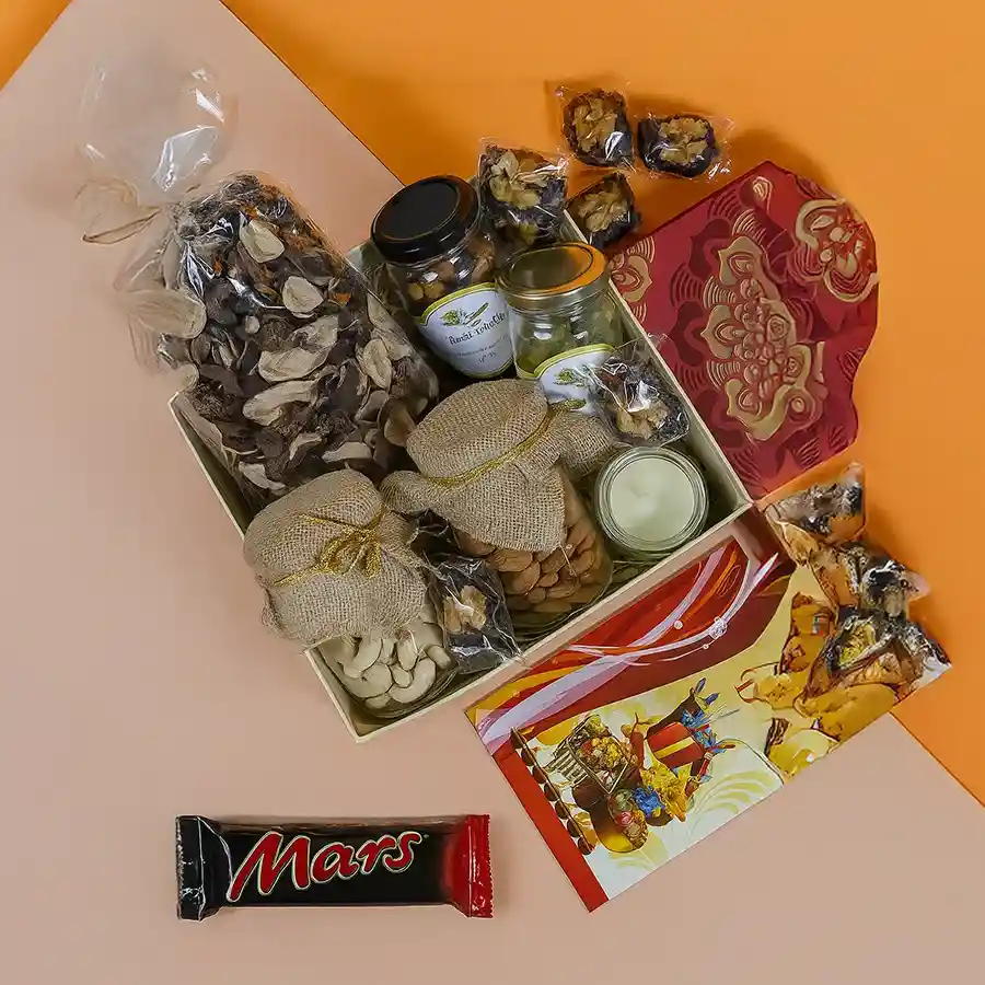 A beautifully arranged Diwali hamper featuring elegant glass jars filled with cashews, almonds, exotic trail mix, walnut-stuffed dates, musk-scented potpourri, and a Bella Vita organic candle, all presented in a reusable Mandala box with a Happy Diwali card.