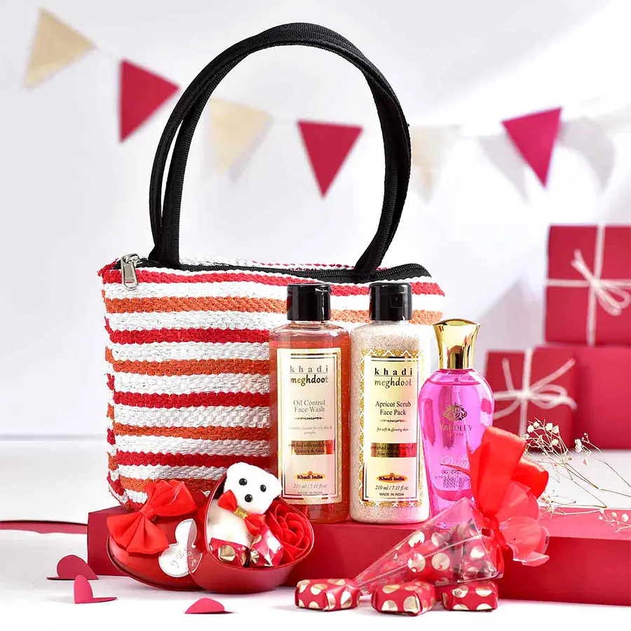 Multicolored Purse N Pamper Hamper featuring a recycled plastic purse, red chocolate cones, a heart tin with chocolates, a teddy, a rose, herbal skincare products, and a perfume.