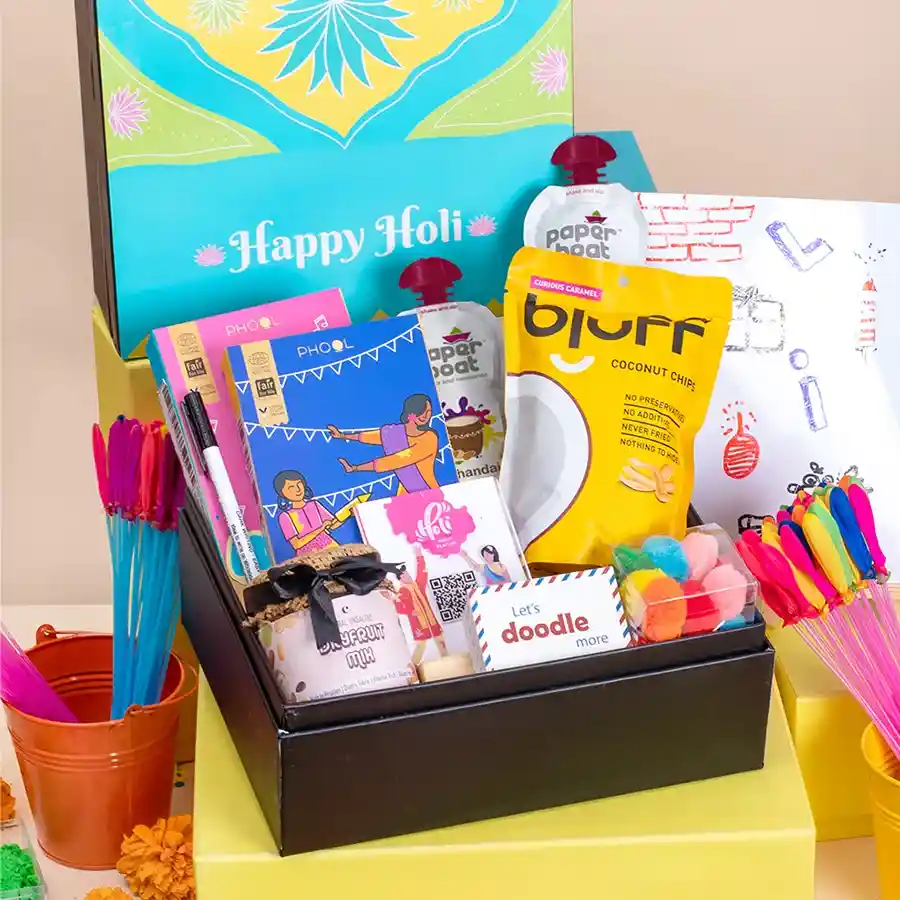 A festive collection of Holi celebration items including colorful powders, snacks, a whiteboard, a Pictionary game, decorative pom poms, and a Holi songs playlist in a stylish reusable box.