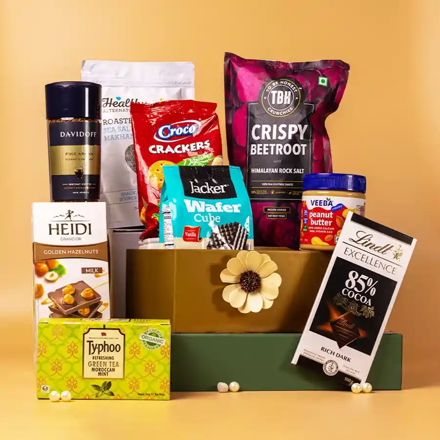 A beautifully arranged gourmet gift hamper filled with assorted treats including peanut butter, crackers, dark chocolate, hazelnut bar, wafer cubes, instant coffee, mint green tea, Makhana pops, and beetroot crunchies, all in a reusable basket.