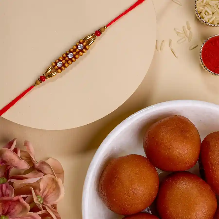 Beautiful AD Rakhi placed next to a box of Gulab Jamun, showcasing the Rakhi Sutra design and a complimentary message card, Roli, Tika, and Chawal for Raksha Bandhan celebration.