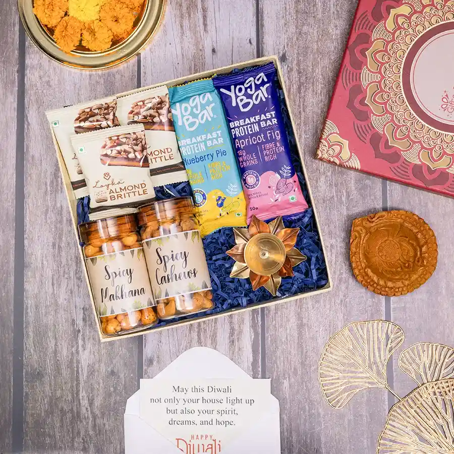 Festive Spice N Light Delight gift box featuring crunchy spicy makhana, cashews, Loyka almond brittle, Yoga Bar apricot fig and blueberry pie bars, a copper lotus diya, and a traditional mud diya, all in a stylish reusable box.